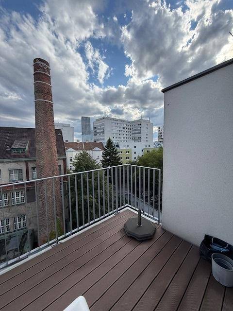 Balkon mit Holzdielen und Metallgeländer bietet einen Blick auf einen alten Schornstein und Stadtgebäude.