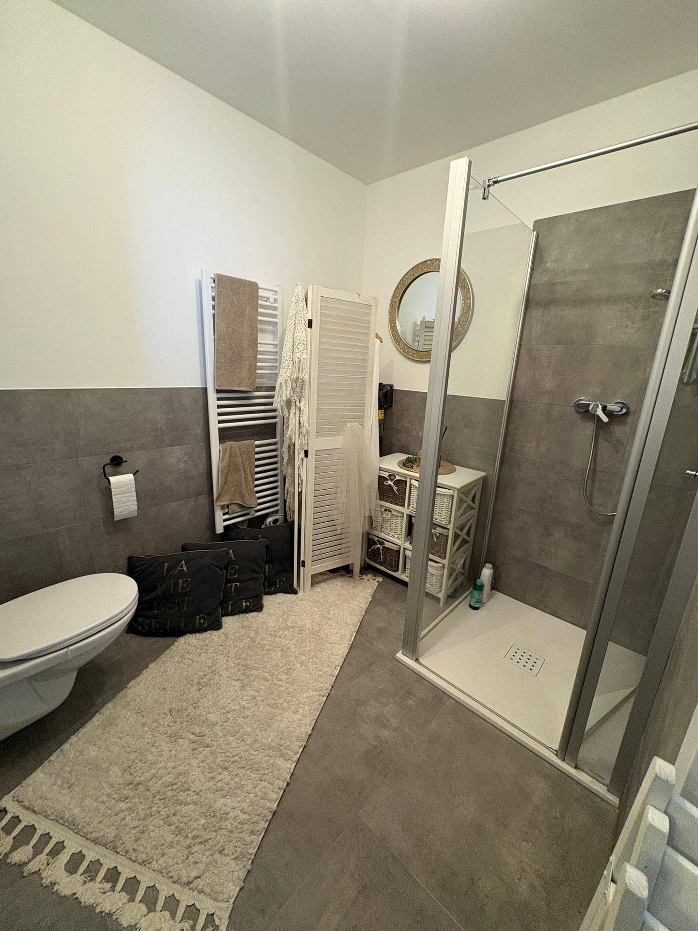 Badezimmer mit grauen Fliesen, wandhängendem WC, Duschkabine und beheiztem Handtuchhalter.