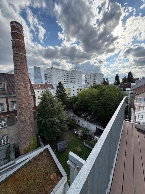 Ausblick von einem Balkon auf einen markanten Backsteinschornstein, grüne Bäume und die Stadtkulisse.