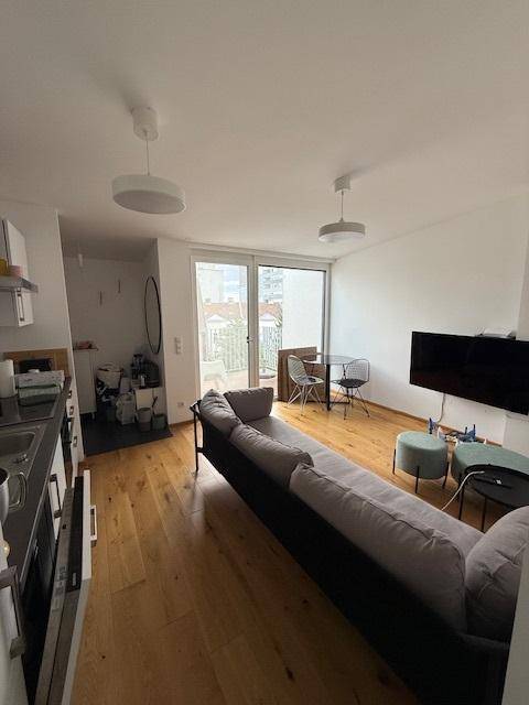 Wohn-Essbereich mit Sofa, Holzboden und Zugang zum Balkon, angrenzend an eine offene Küche.