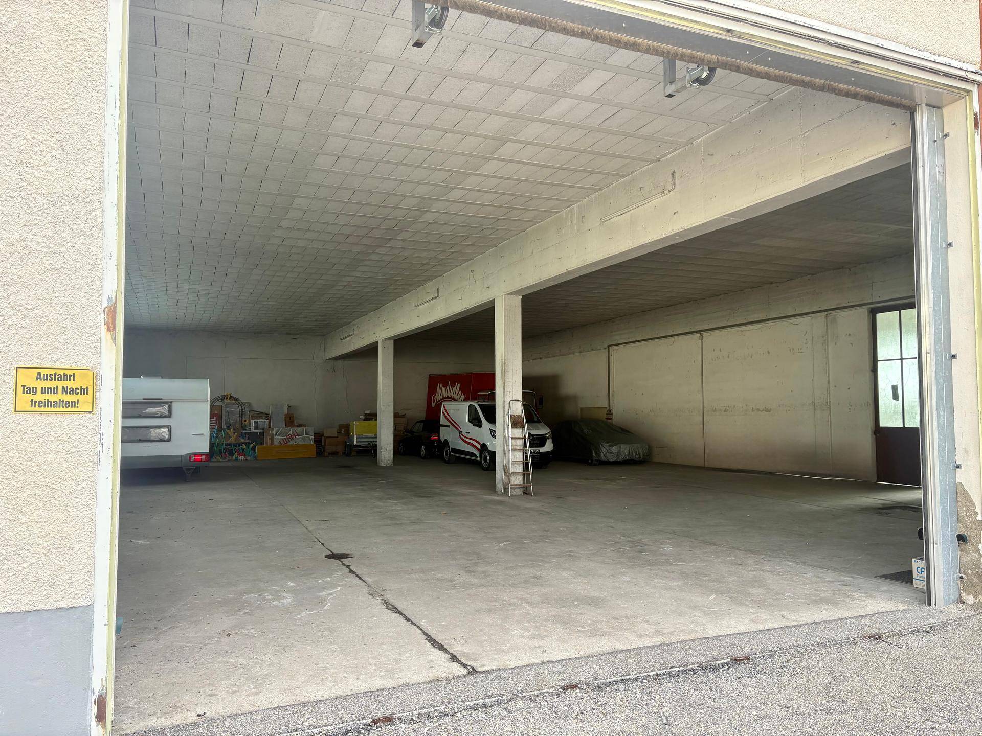 Weitläufige Lagerhalle mit großem Rolltor und Betonboden, bietet viel Platz für Fahrzeuge und Güter.