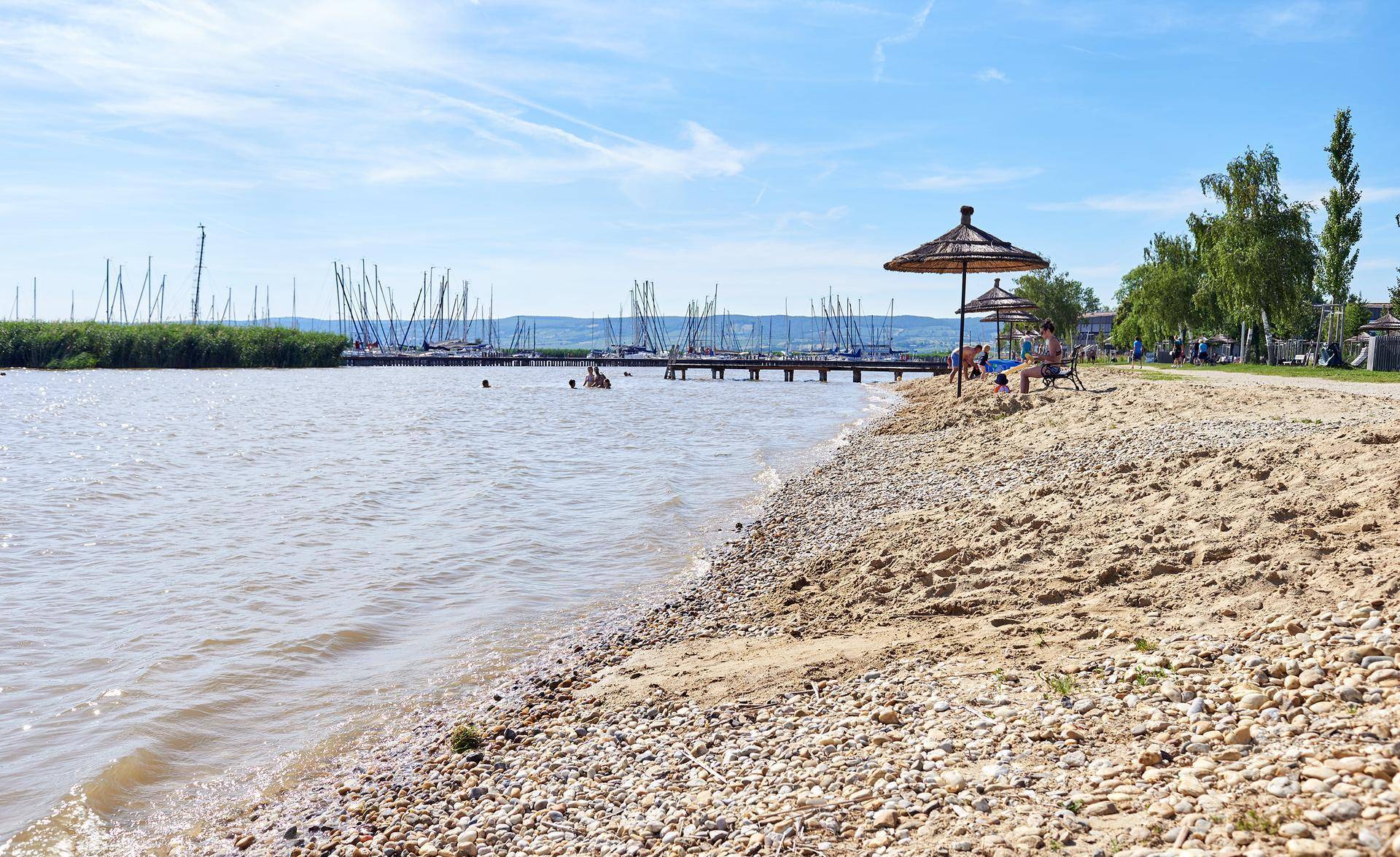 Kiesstrand an einem See mit Sonnenschirmen und Booten im Hintergrund unter blauem Himmel.