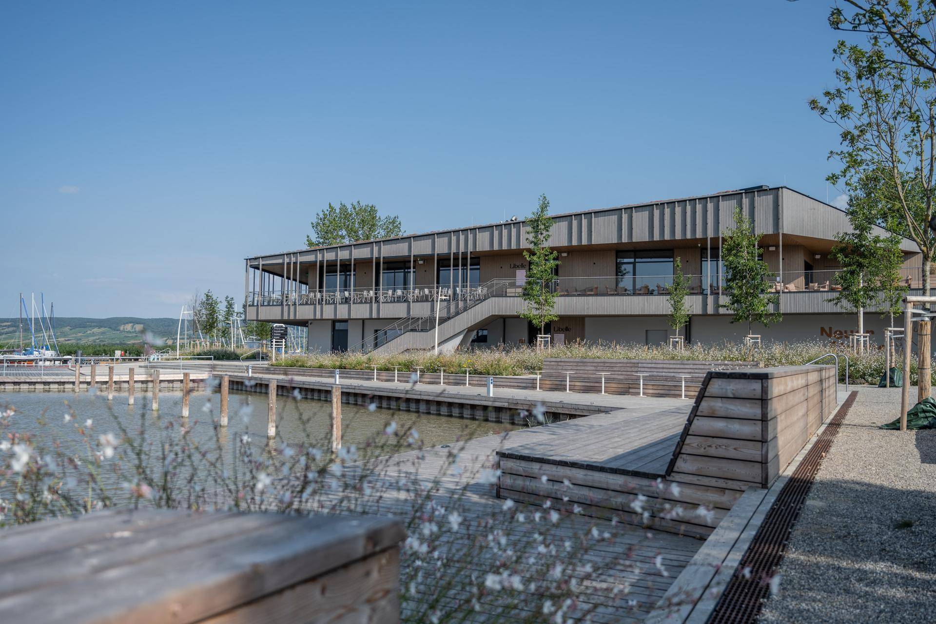 Modernes Gebäude mit Holzstegen am Wasser, umgeben von Natur und klarem Himmel.