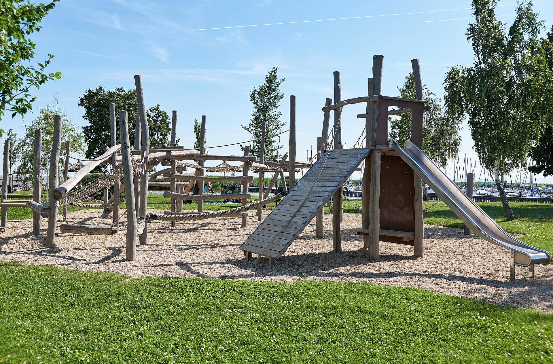 Großer Holzspielplatz mit Rutsche und Klettermöglichkeiten auf Sandboden, umgeben von Rasen und Bäumen.