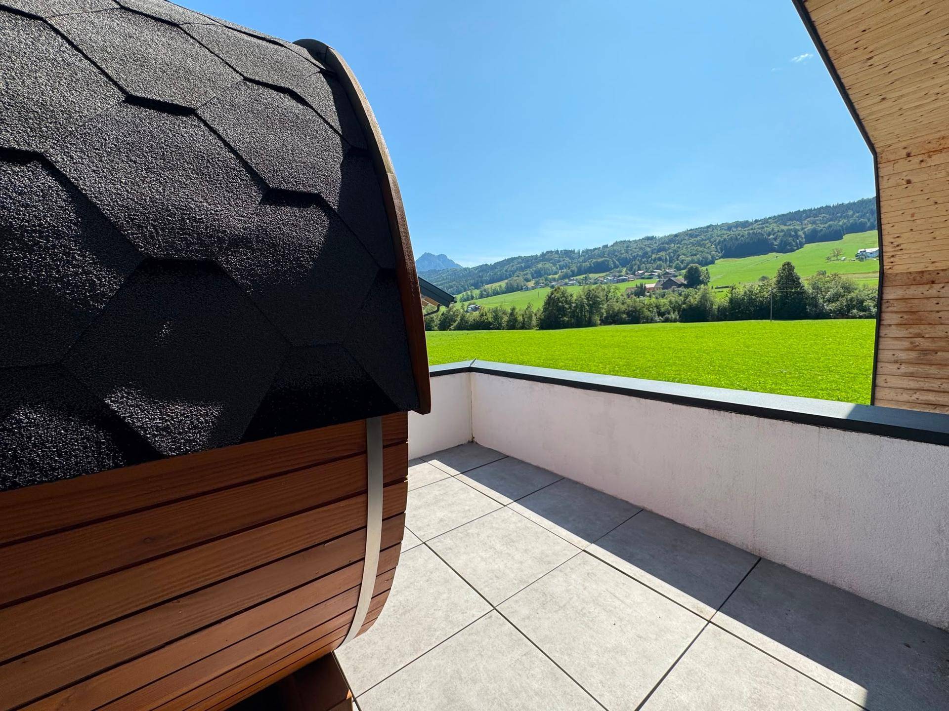 Sonnenterrasse mit Fasssauna und weitem Blick auf die grüne Berglandschaft.