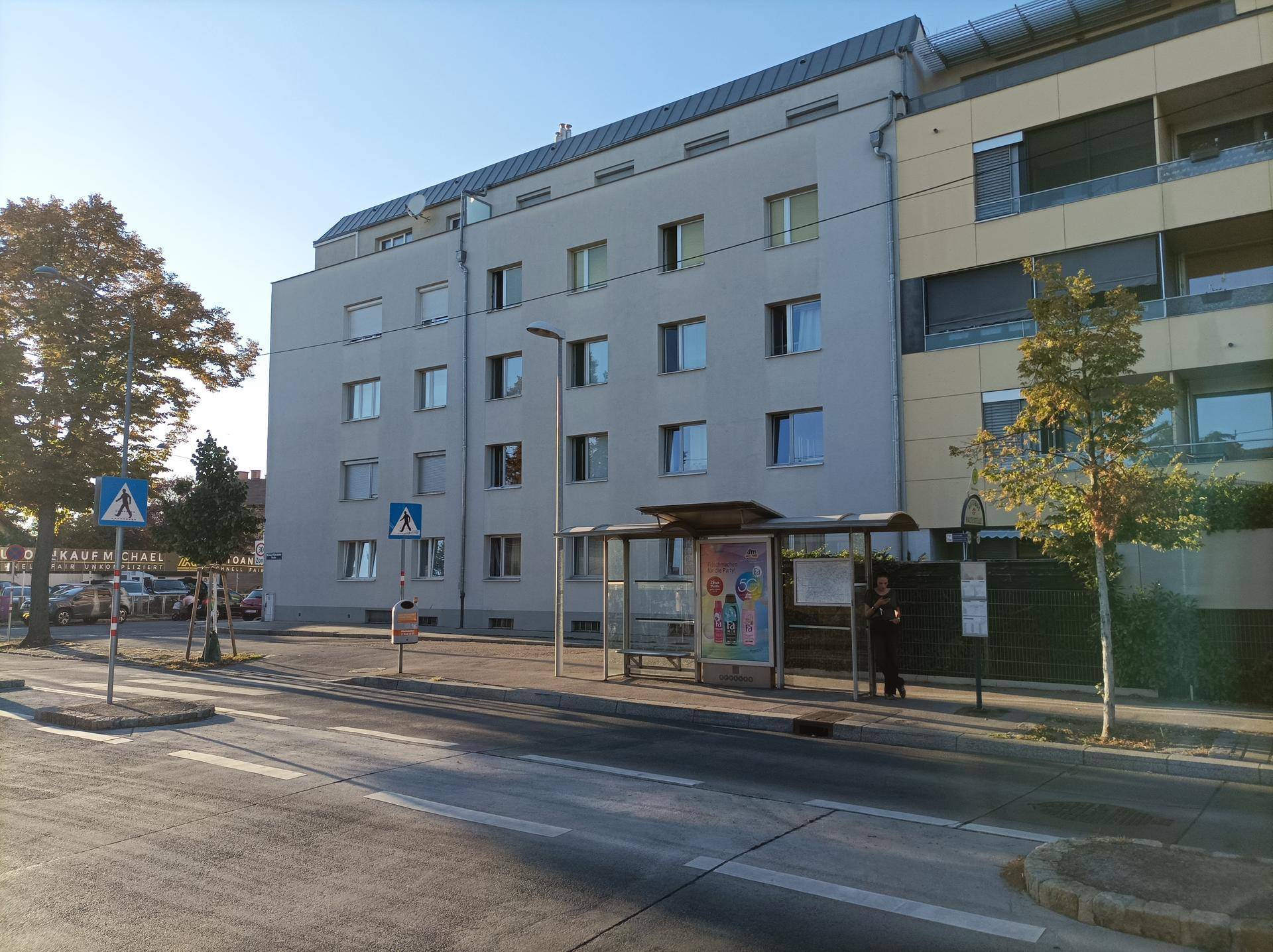 Modernes Mehrfamilienhaus an einer Straße mit Bushaltestelle und Bäumen in städtischer Umgebung.