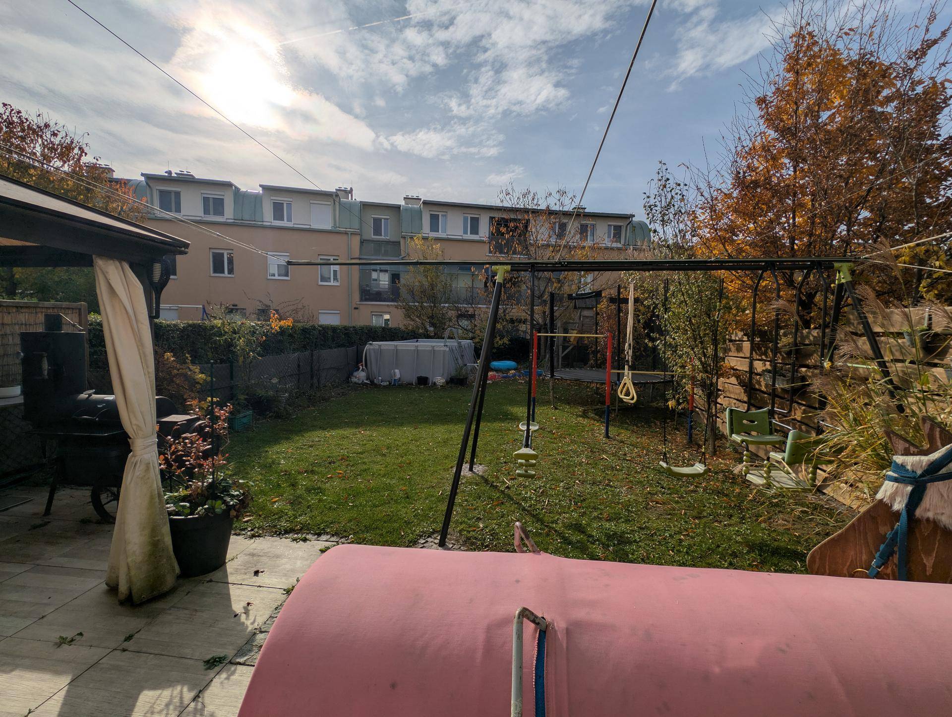 Sonniger Garten mit Rasenfläche und Spielgeräten, umgeben von Hecken und der Rückseite des Gebäudes.