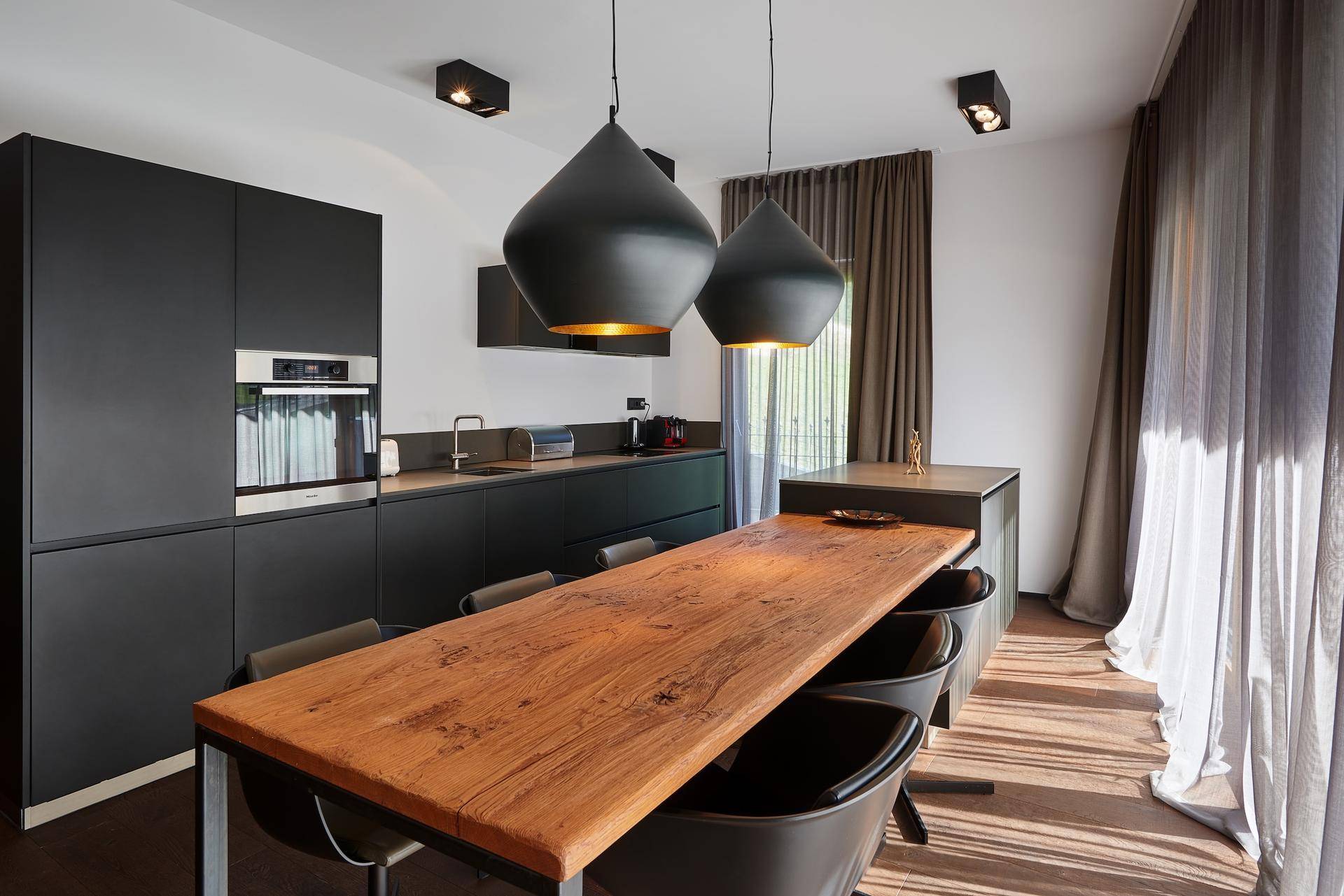 Elegante schwarze Küche mit Holztisch und modernen Pendelleuchten, viel Tageslicht.