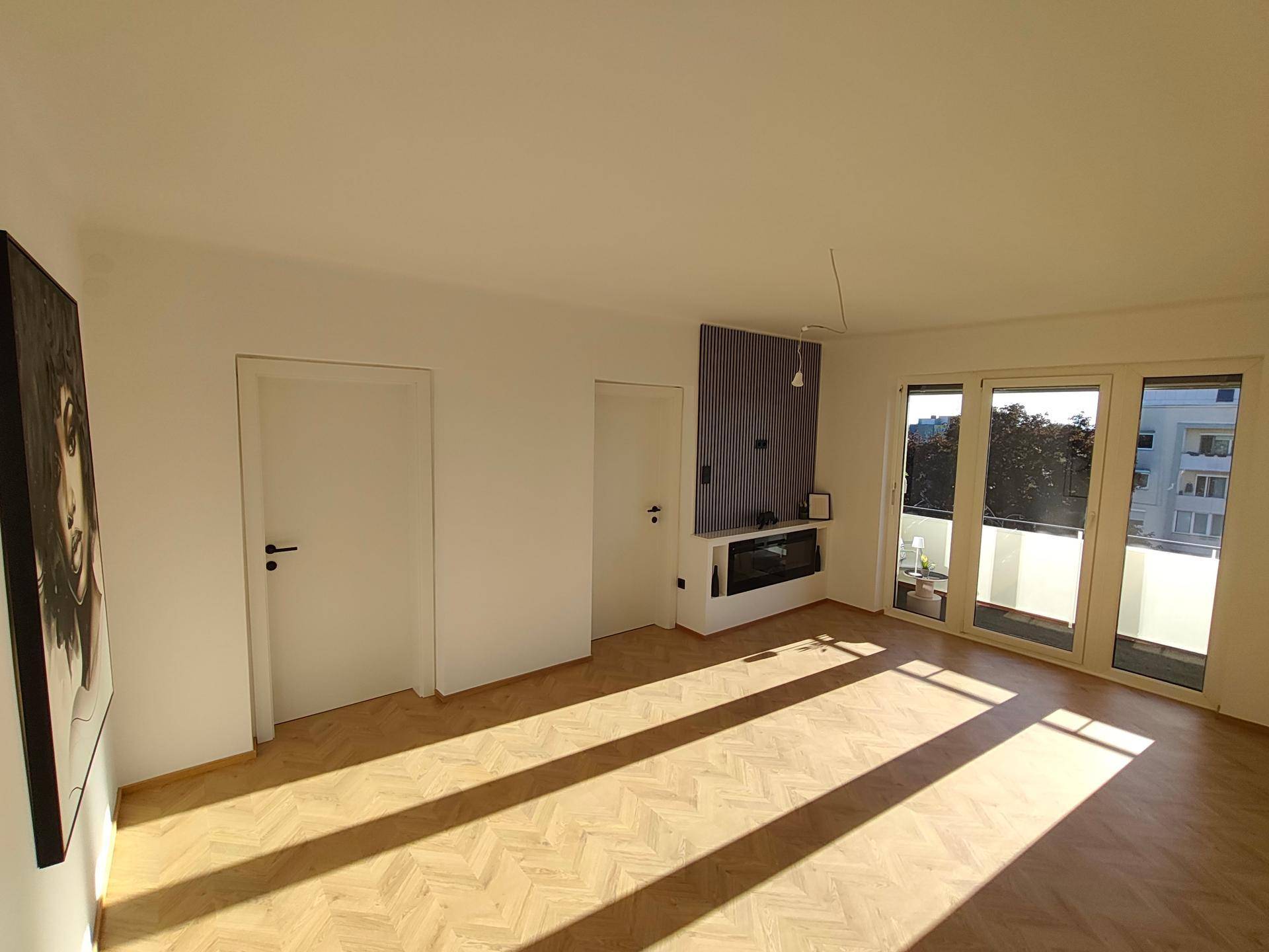 Geräumiges Wohnzimmer mit Elektrokamin, Fischgrätparkett und Zugang zu einem sonnigen Balkon.