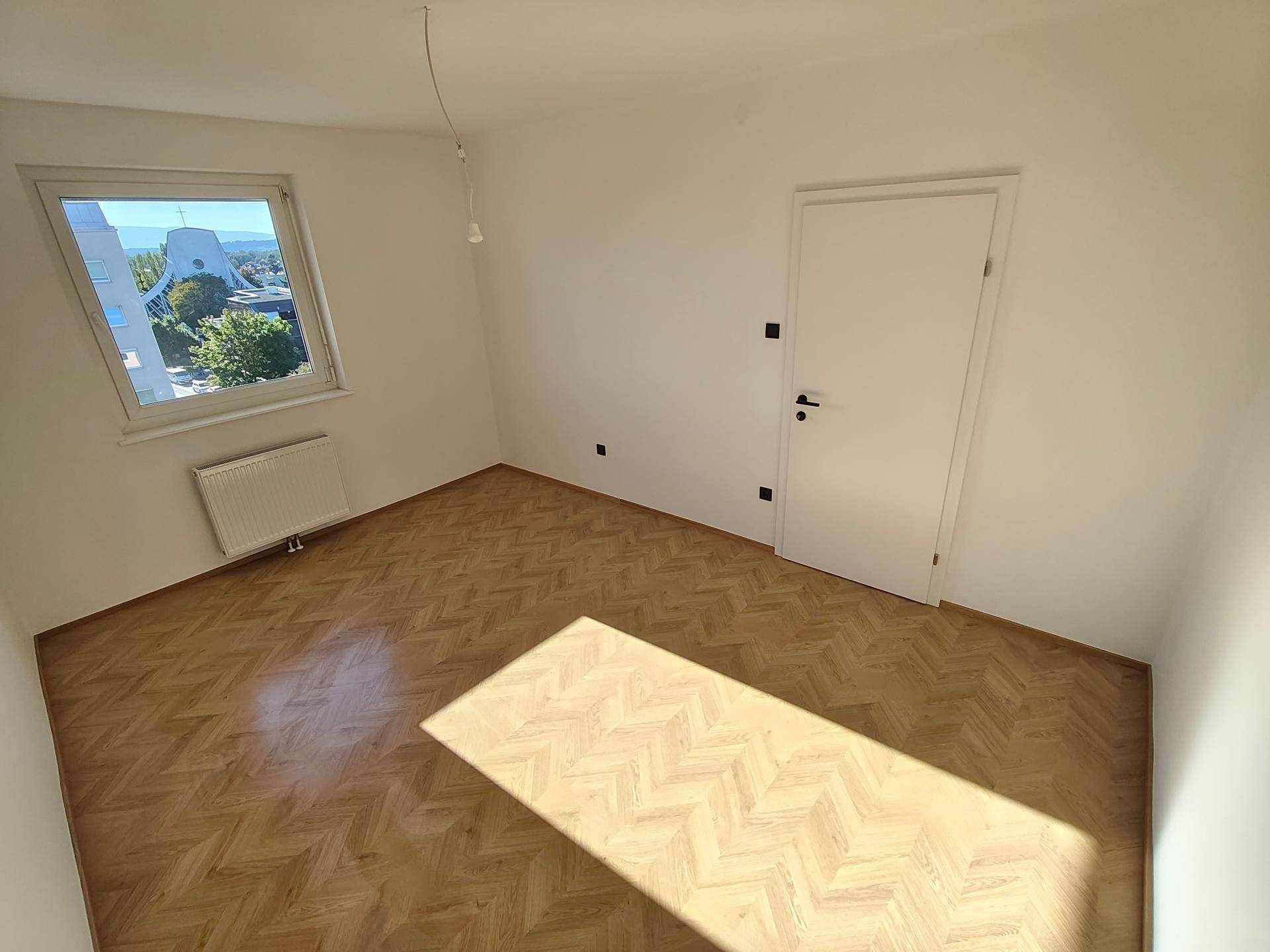 Helles Schlafzimmer mit Fischgrätparkett, weißen Wänden und Fenster mit Ausblick.