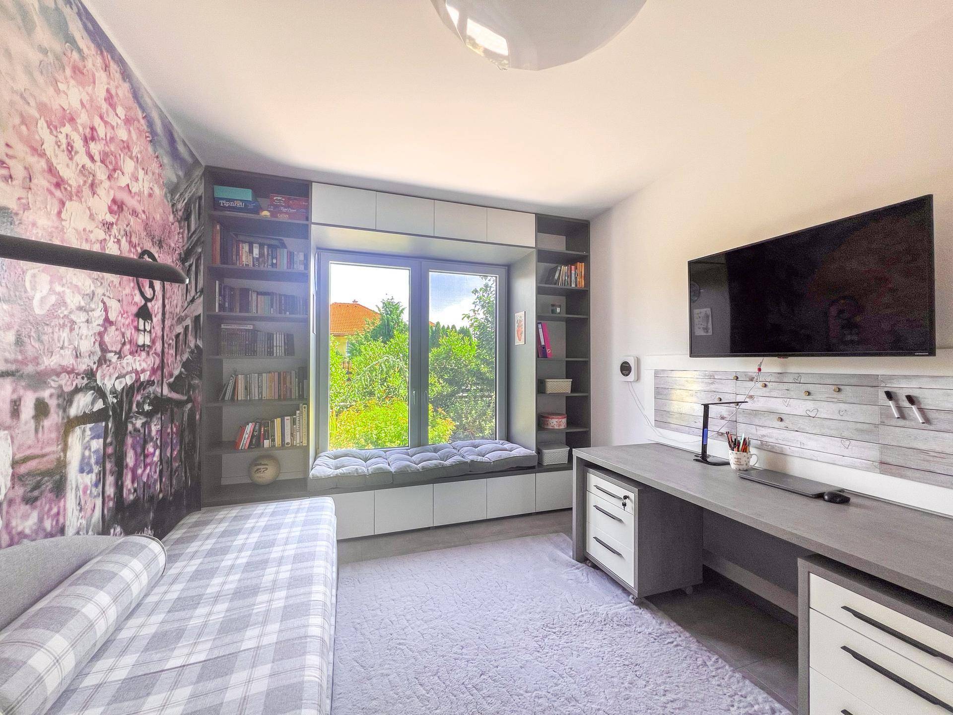 Schlafzimmer mit Schreibtisch, integrierten Regalen und großem Fenster mit Blick ins Grüne.