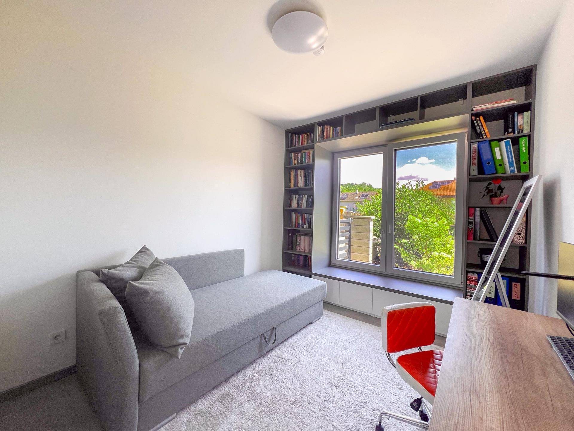 Helles Zimmer mit Schlafsofa, Schreibtisch und Bücherregalen um das Fenster herum.