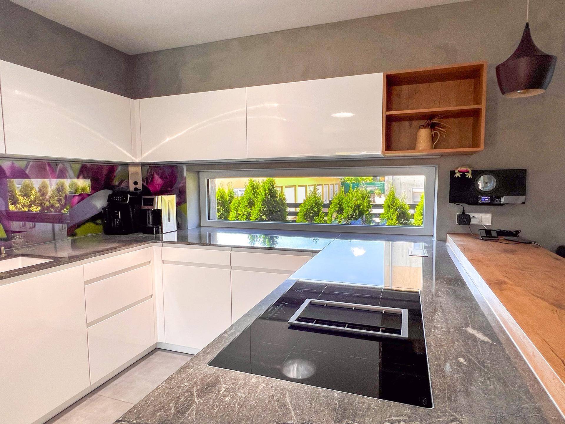 Moderne Küche mit weißen Hochglanzfronten, Induktionskochfeld und Fenster mit Blick ins Grüne.