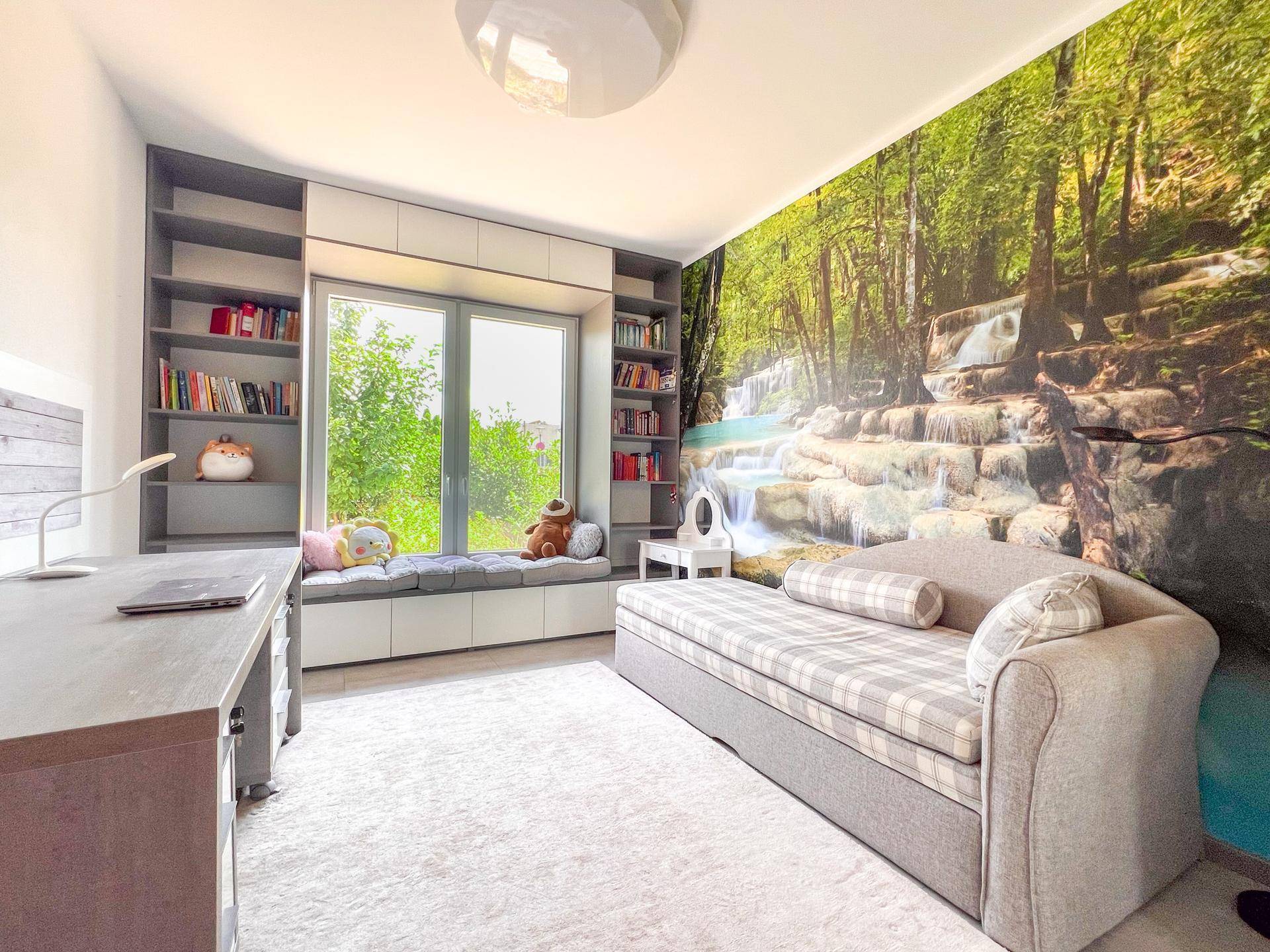 Helles Zimmer mit Schreibtisch, integrierten Regalen und Wandtapete mit Wasserfallmotiv.