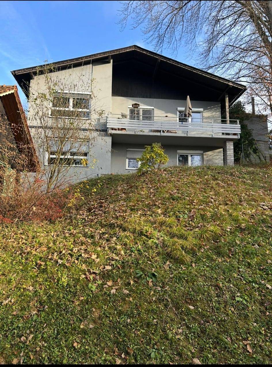 Gesamtansicht des Hauses mit grauer Fassade, Balkon und umgebender Natur im Herbst.
