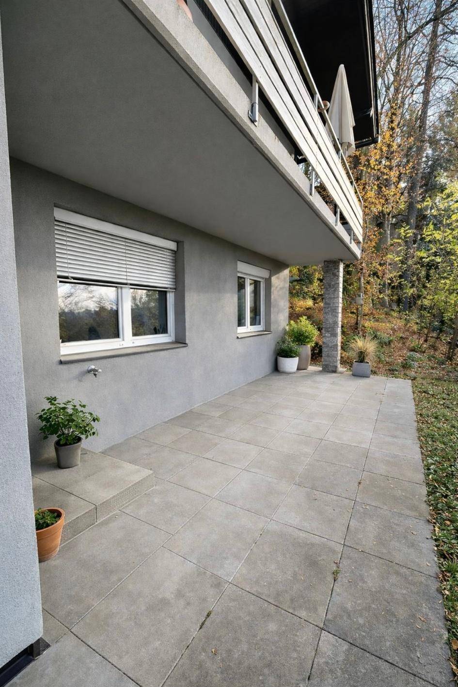 Gepflasterte Terrasse im Erdgeschoss mit Blick ins Grüne und einem darüberliegenden Balkon.