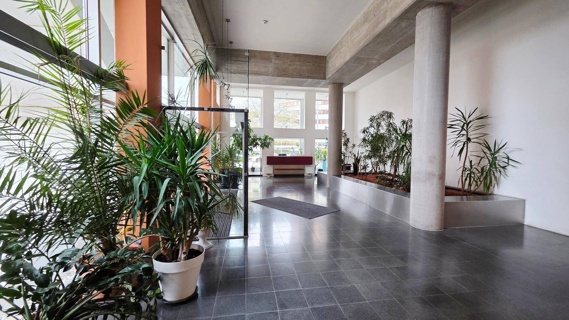 Geräumige moderne Lobby mit großen Fenstern, viel Grün und Fliesenboden.
