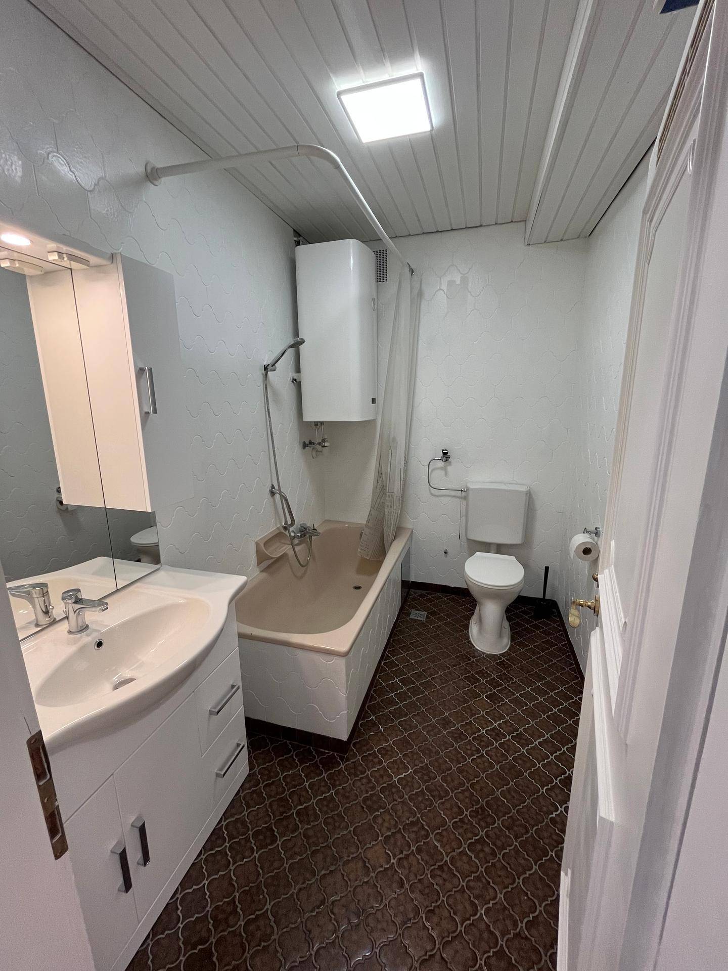 Badezimmer mit Badewanne, WC, Waschbecken und Spiegelschrank, ausgestattet mit dunklen Fliesen.