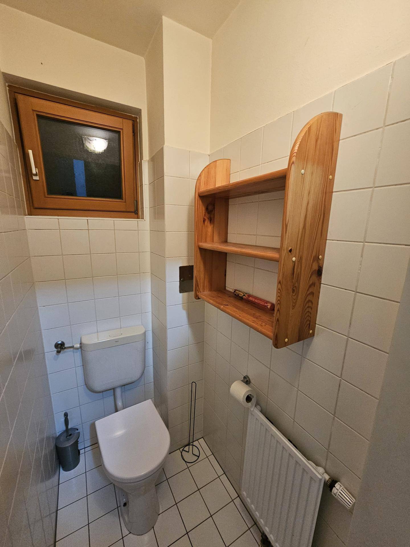 Kleines WC mit Fenster, Toilette und einem praktischen Wandregal aus Holz.