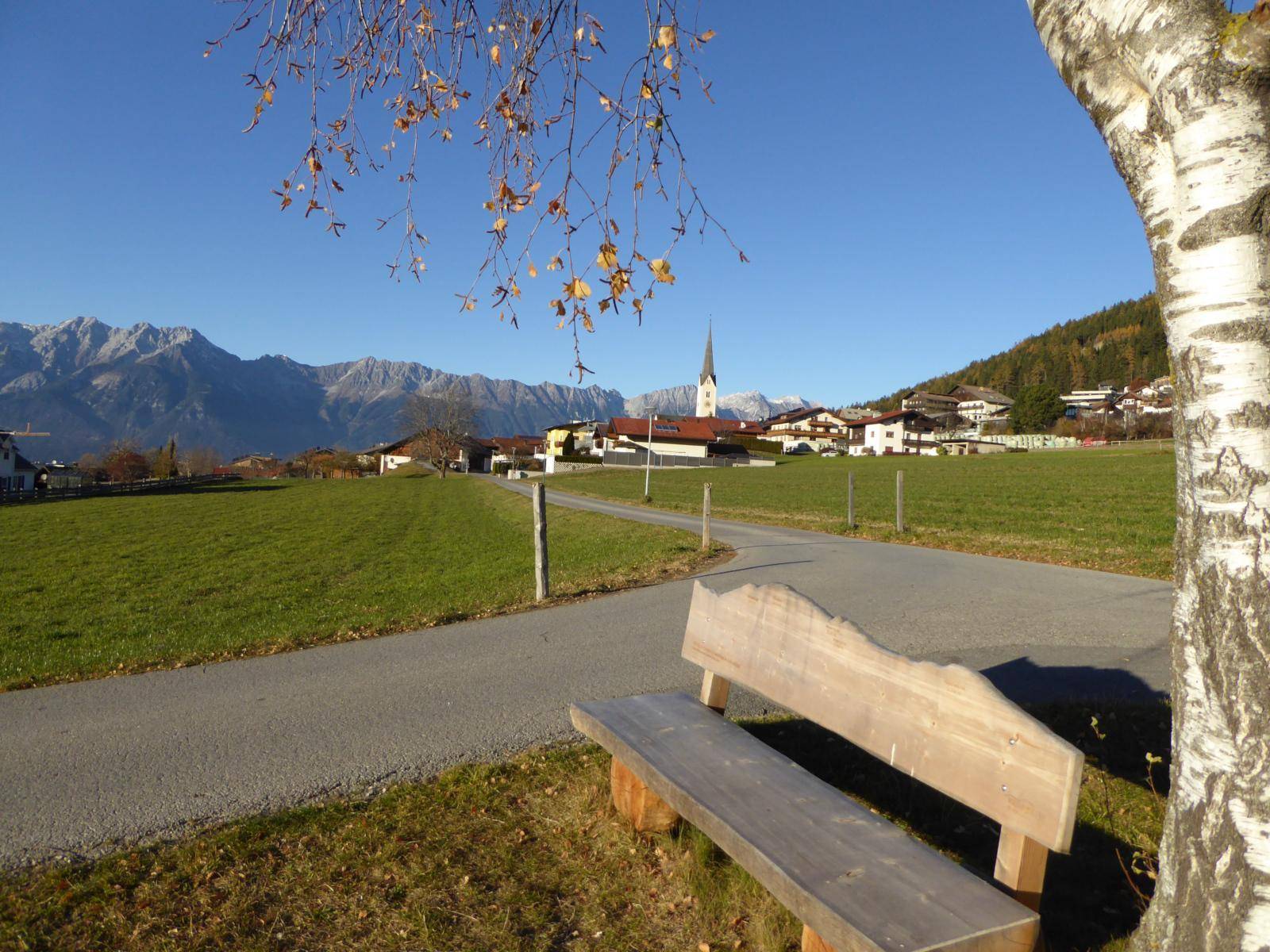 Idyllischer Landschaftsblick mit einem Weg, einer Holzbank und einem Dorf mit Kirchturm vor Bergkulisse.