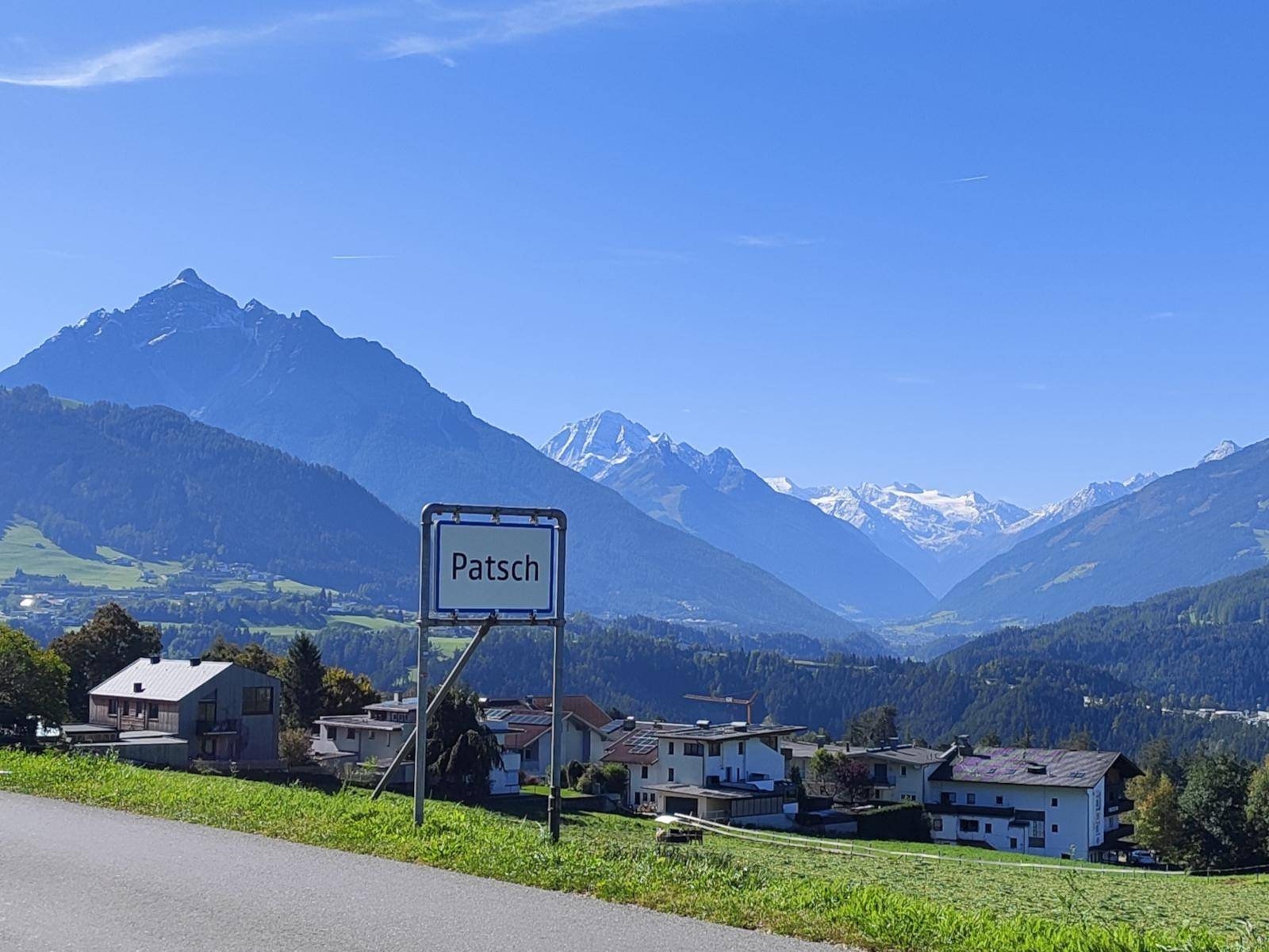 Panoramablick auf das Dorf Patsch mit majestätischen Bergen im Hintergrund unter blauem Himmel.