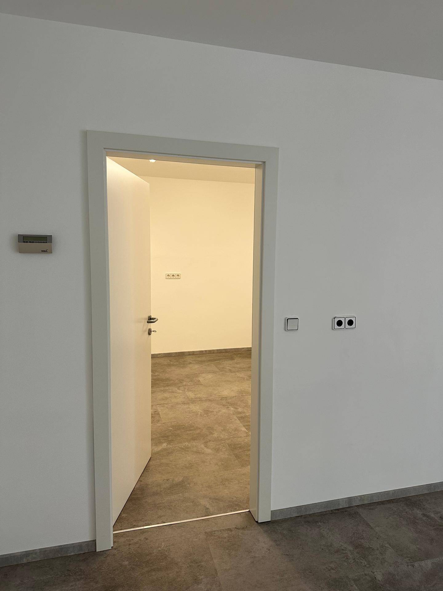 Offene weiße Tür führt in einen weiteren leeren Raum mit Fliesenboden, weißen Wänden und sichtbaren Steckdosen.