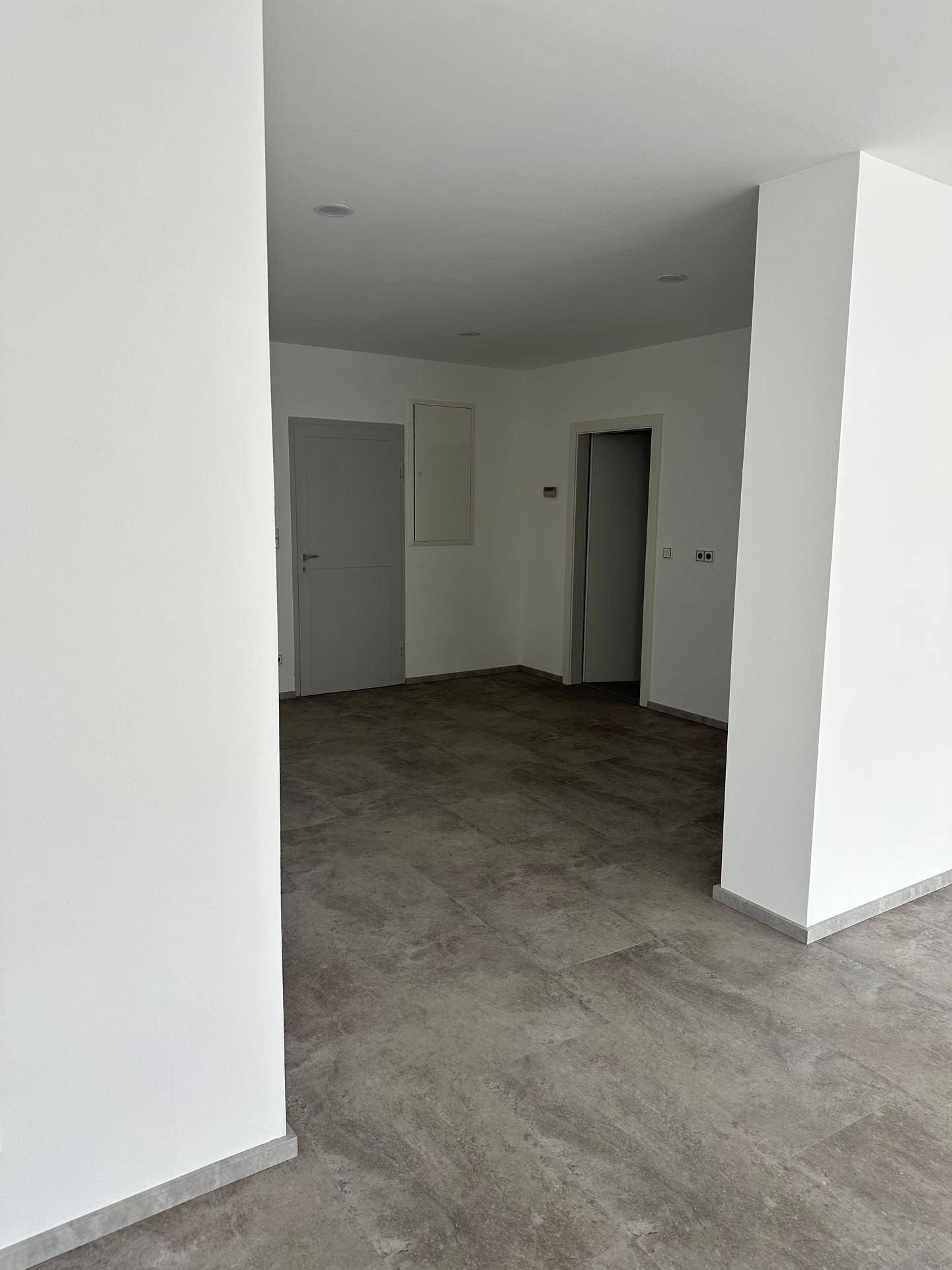 Leerer Innenraum mit grauem Fliesenboden und weißen Wänden, der Zugang zu mehreren weiteren Räumen bietet.