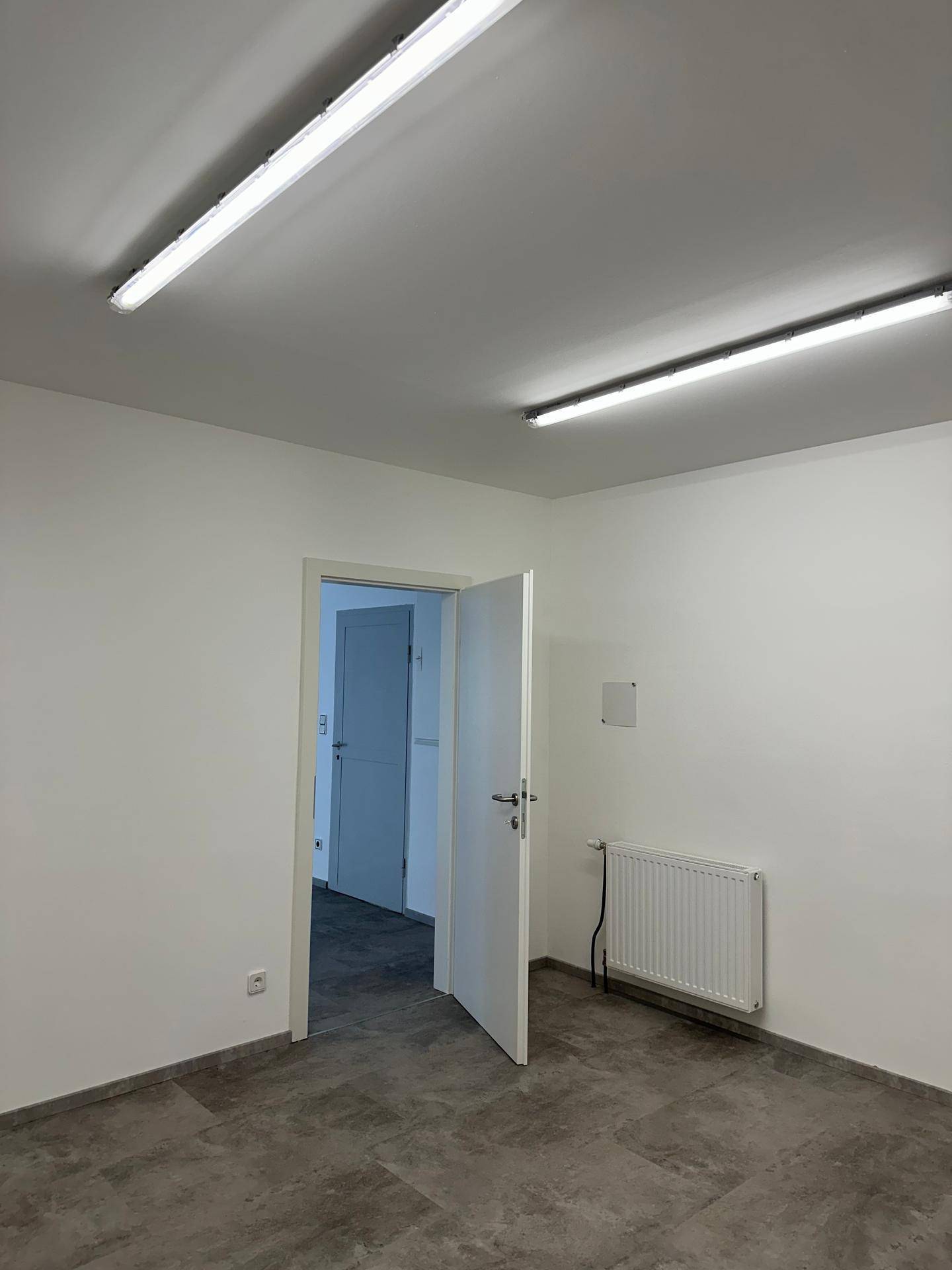 Leerer Raum mit Fliesenboden, weißen Wänden, Heizkörper und Deckenleuchten, bereit für die individuelle Gestaltung.