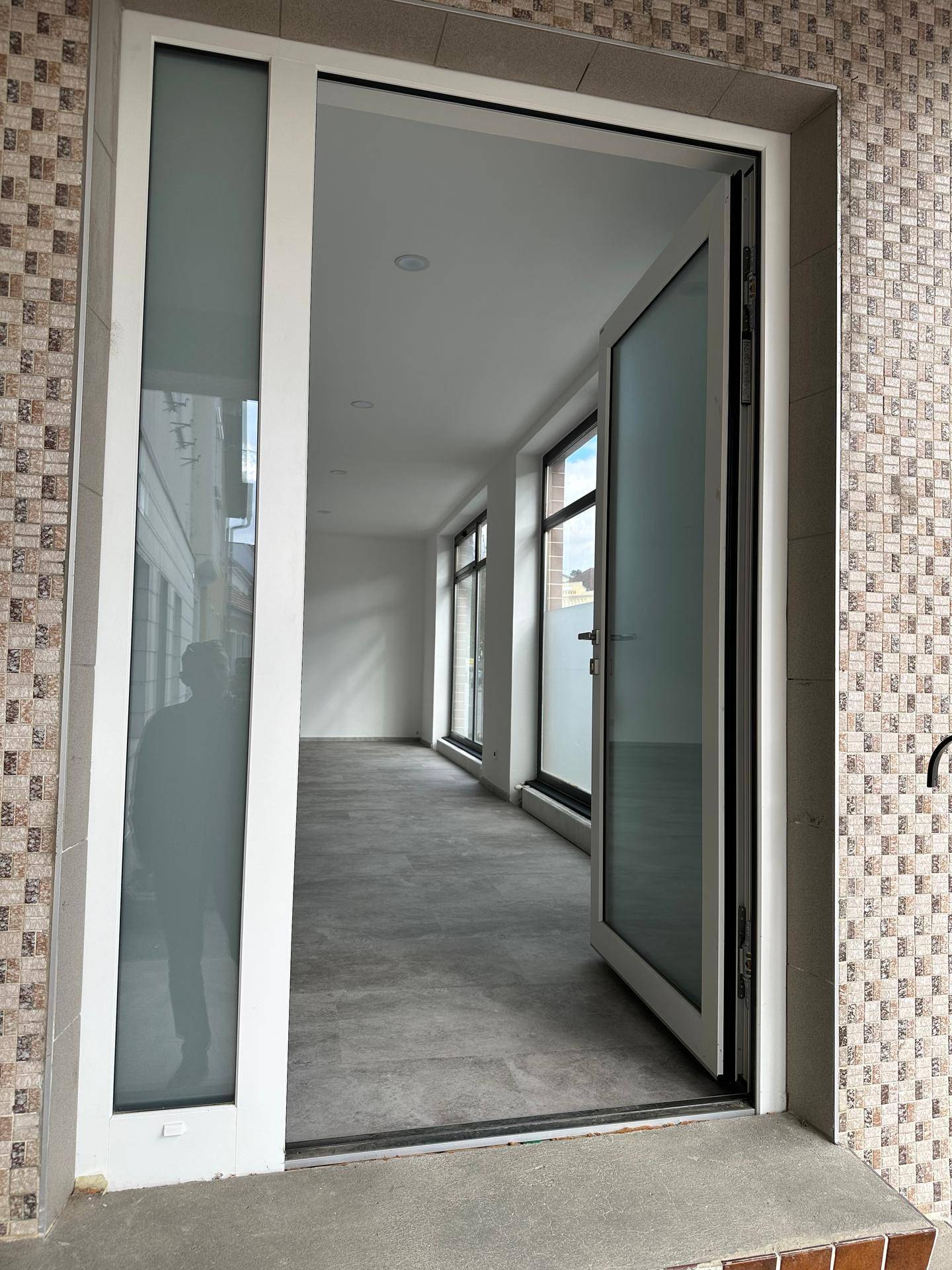 Blick durch die offene Glastür in einen leeren, hellen Geschäftsraum mit großen Fenstern und modernem Fliesenboden.