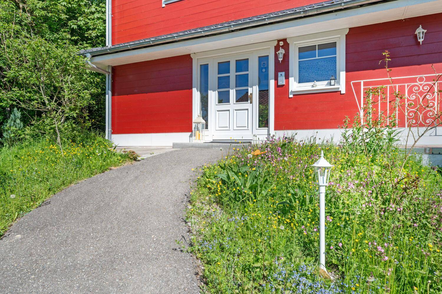 Eingangsbereich des roten Hauses mit gepflastertem Weg und blühenden Blumenbeeten.