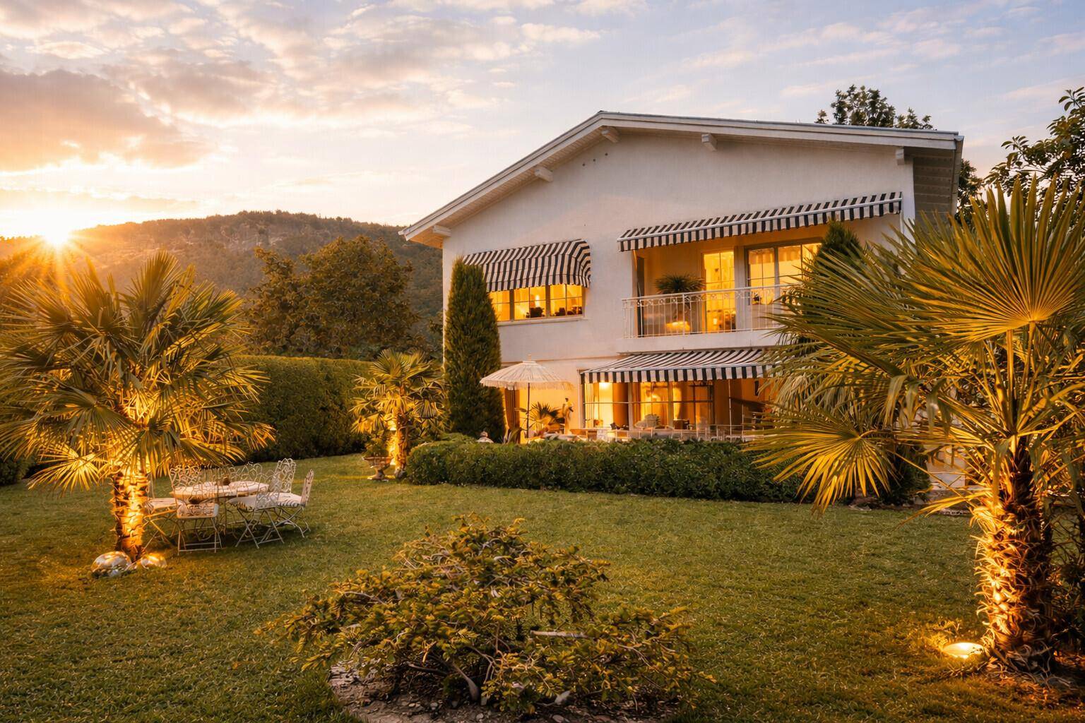 Das Haus und der Garten in der Abendsonne mit beleuchteten Palmen und gemütlichen Sitzgelegenheiten.