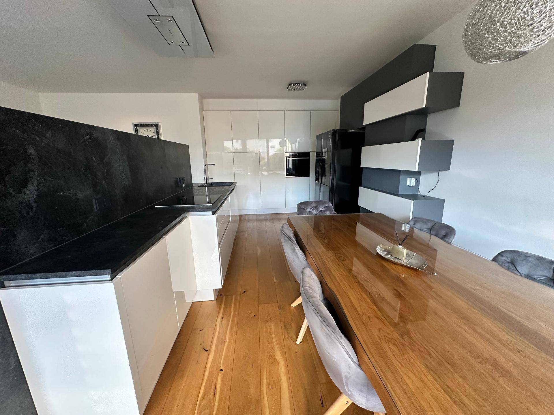 Moderne Küche mit weißen Hochglanzfronten, dunklen Arbeitsplatten und großem Holzesstisch.