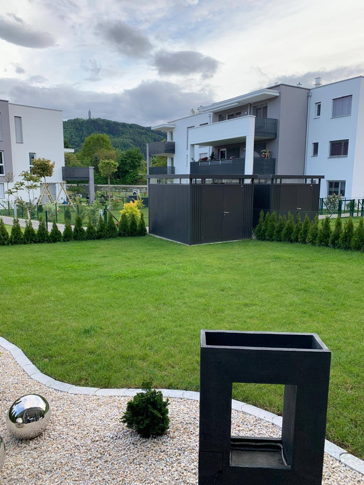 Gepflegter Gemeinschaftsgarten mit Rasenfläche und Blick auf moderne Wohngebäude und Hügel.