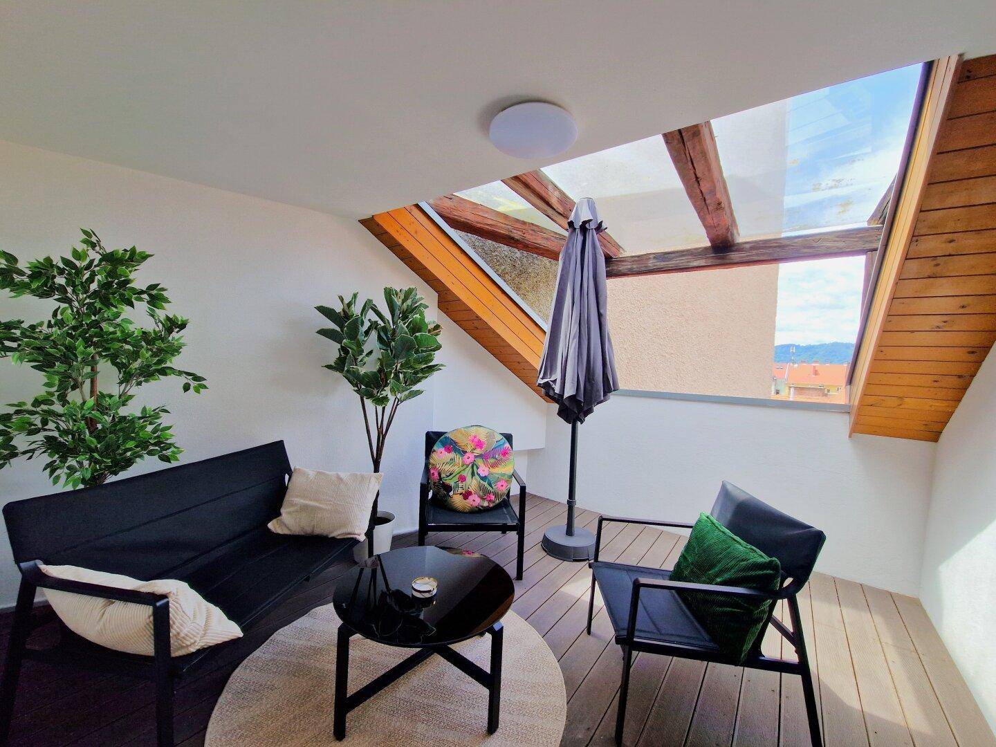Gemütliche Dachterrasse mit Sitzgelegenheiten, Sonnenschirm und Holzbalken unter einem Glasdach.