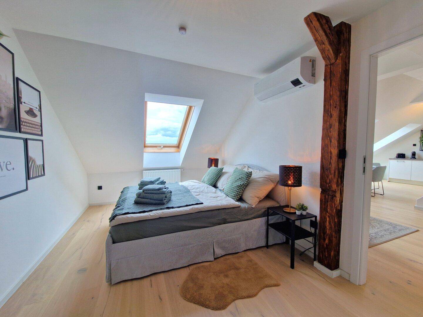 Helles Schlafzimmer mit Dachschräge, Holzbalken, Doppelbett und Dachfenster für natürliches Licht.