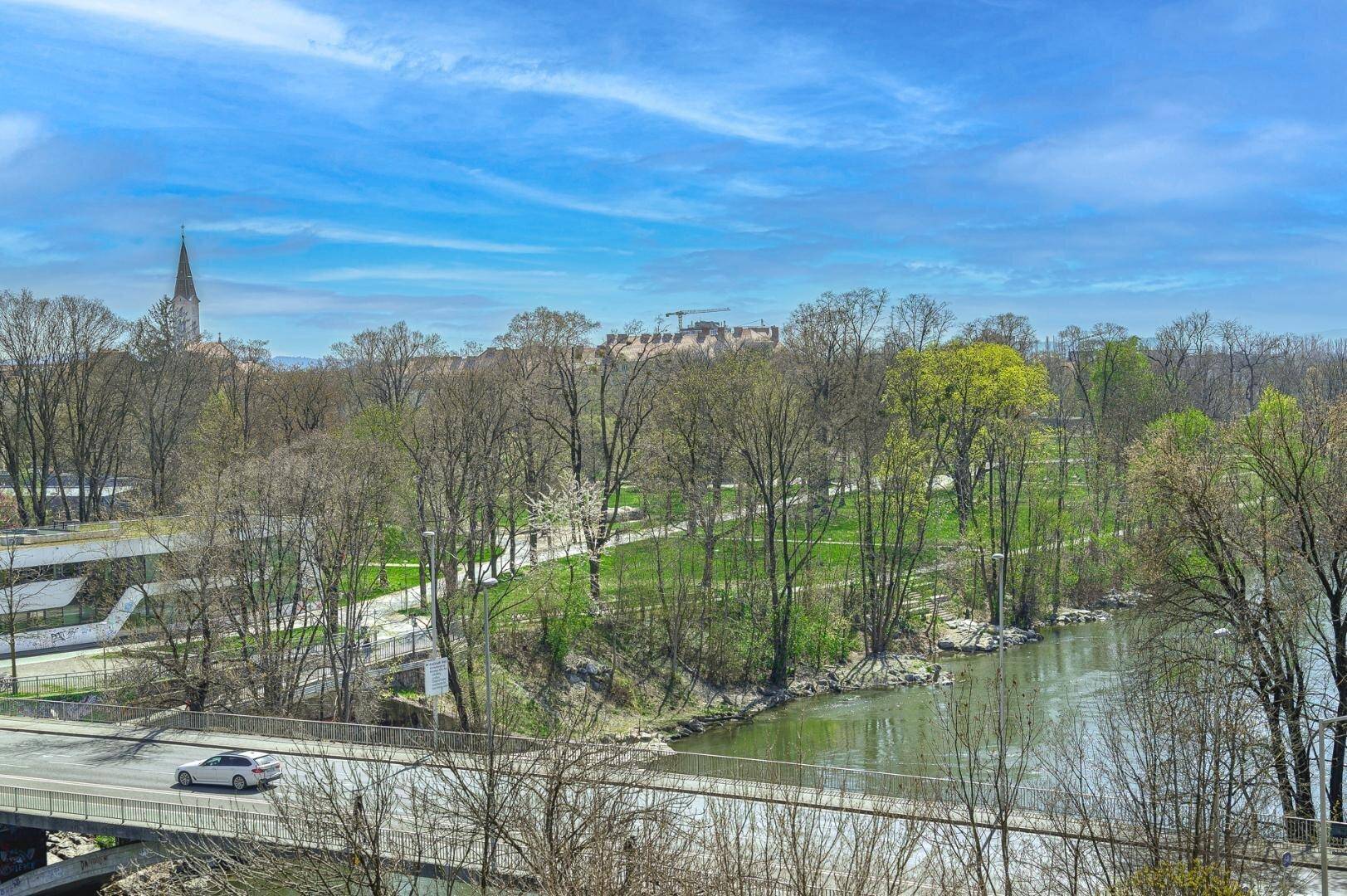 Weitläufiger Blick auf einen Fluss, grüne Bäume und eine Stadtlandschaft unter blauem Himmel.