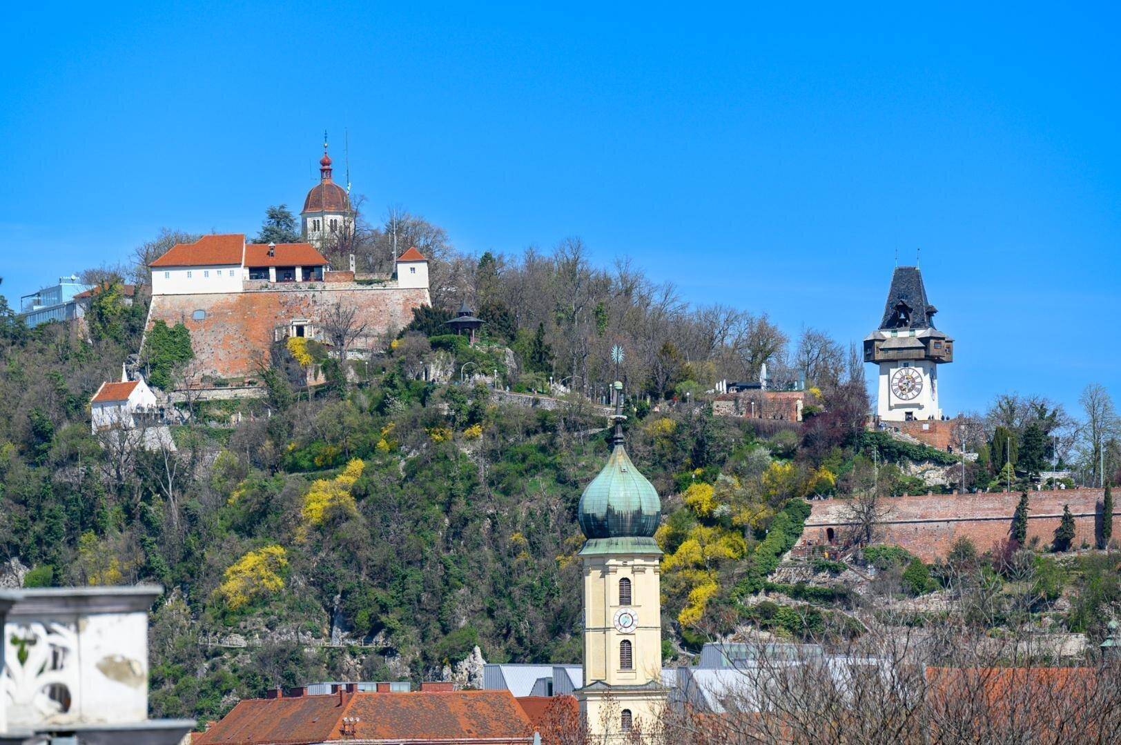 Panoramablick auf die Grazer Altstadt mit Uhrturm und Schlossberg unter strahlend blauem Himmel.