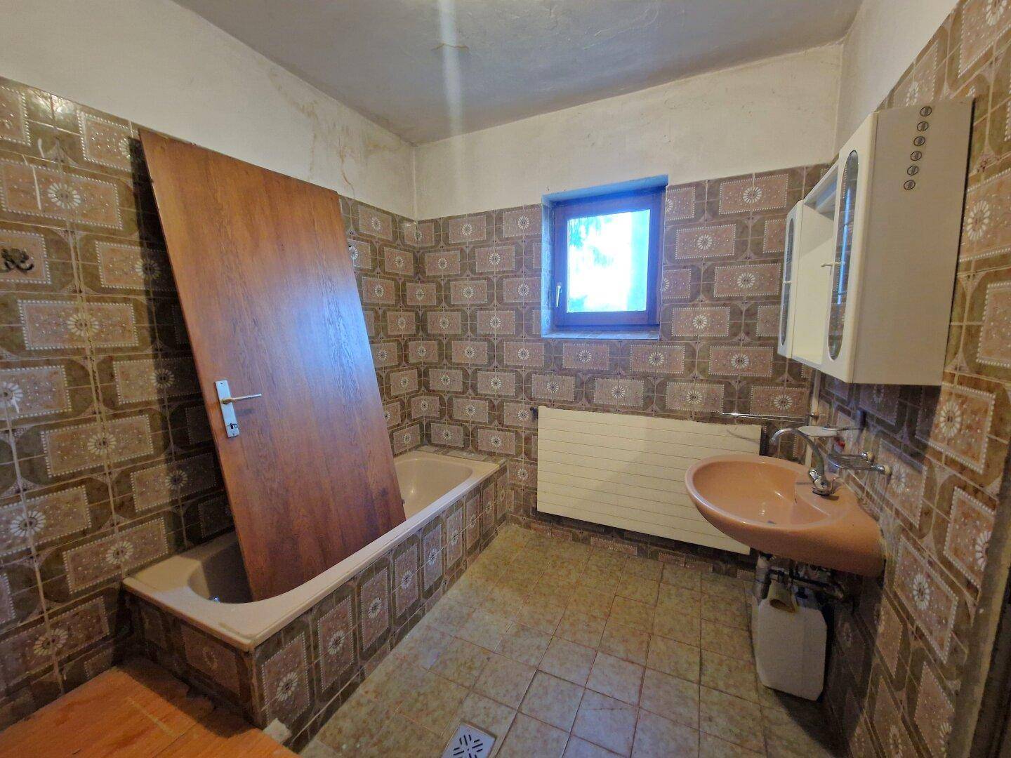 Badezimmer mit Badewanne, Waschbecken und Fenster, das eine umfassende Modernisierung benötigt.