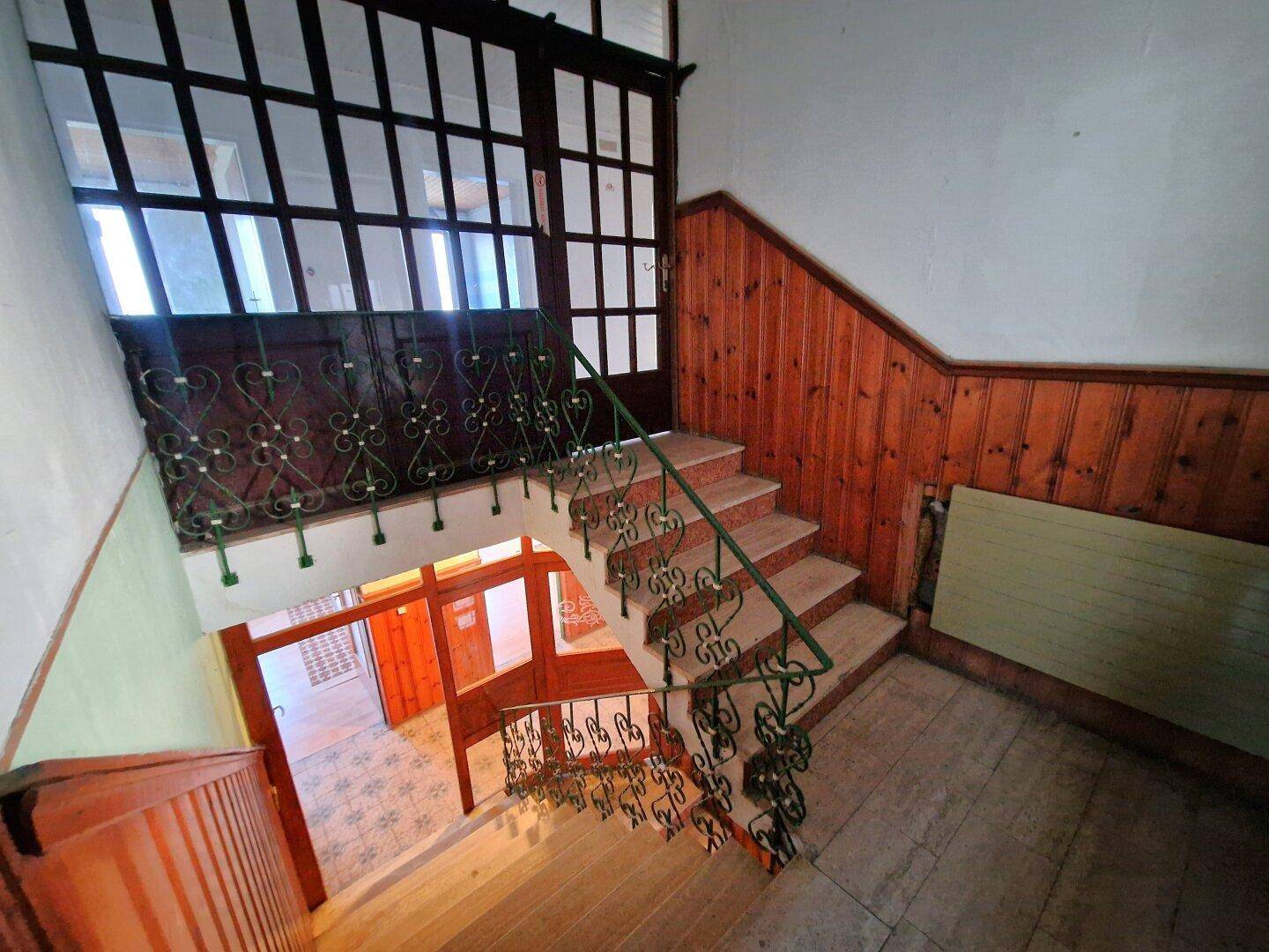 Treppenhaus mit Holzstufen, schmiedeeisernem Geländer und Blick auf den Eingangsbereich.