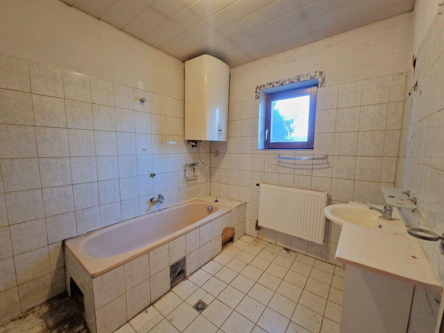 Badezimmer mit Badewanne, Waschbecken und Fenster, das eine umfassende Modernisierung benötigt.