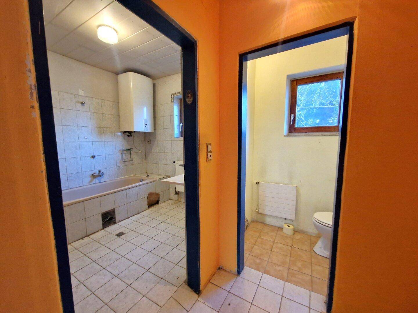 Blick auf ein Badezimmer mit Badewanne und ein separates WC, beide in sanierungsbedürftigem Zustand.