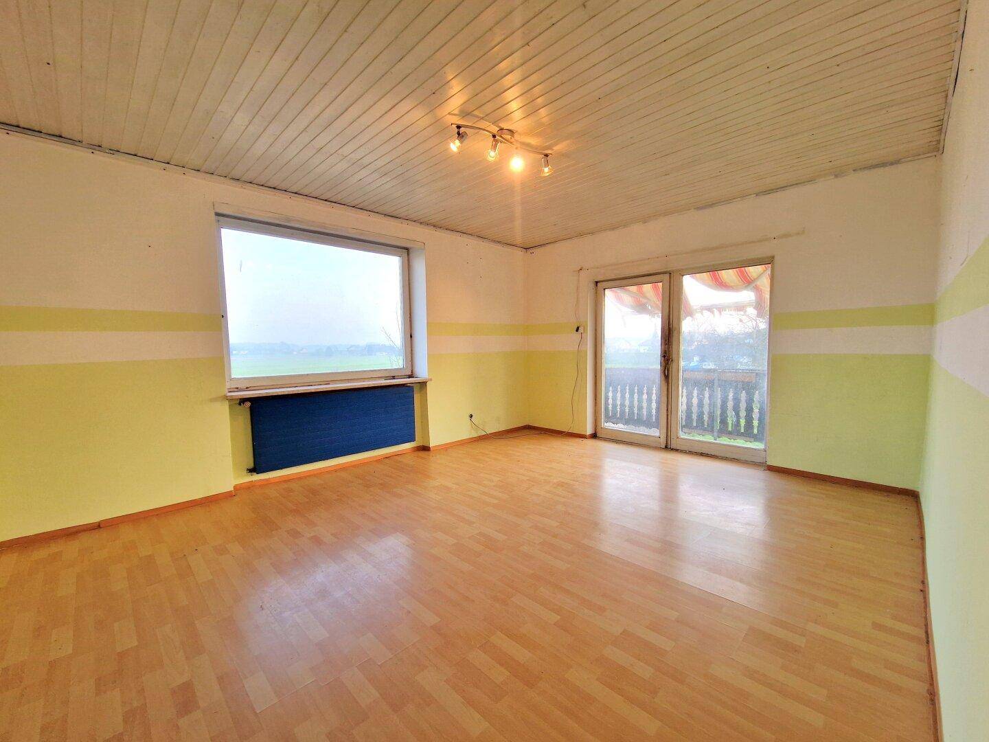 Heller Raum mit Holzbalkendecke, Parkettboden und Zugang zu einem Balkon mit Blick ins Grüne.