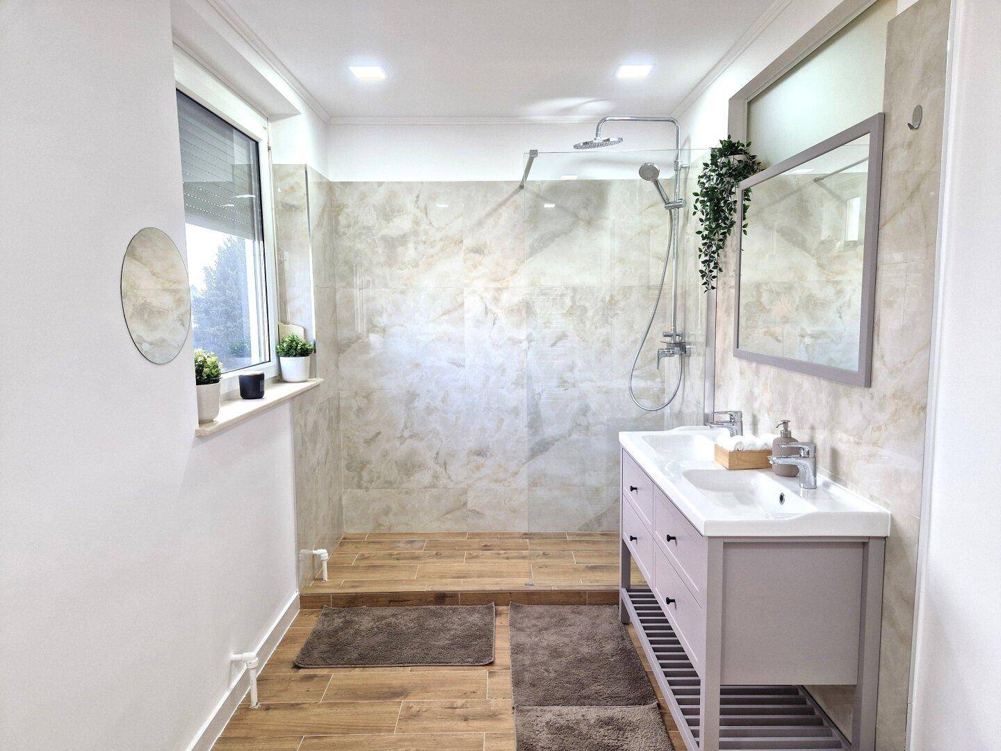 Helles Badezimmer mit Fenster, begehbarer Dusche und Doppelwaschbecken.