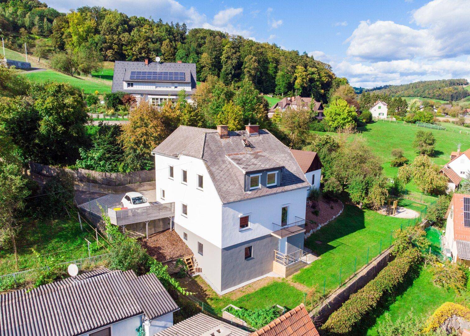 Luftaufnahme eines Einfamilienhauses mit Satteldach, umgeben von viel Grün und Hügellandschaft.
