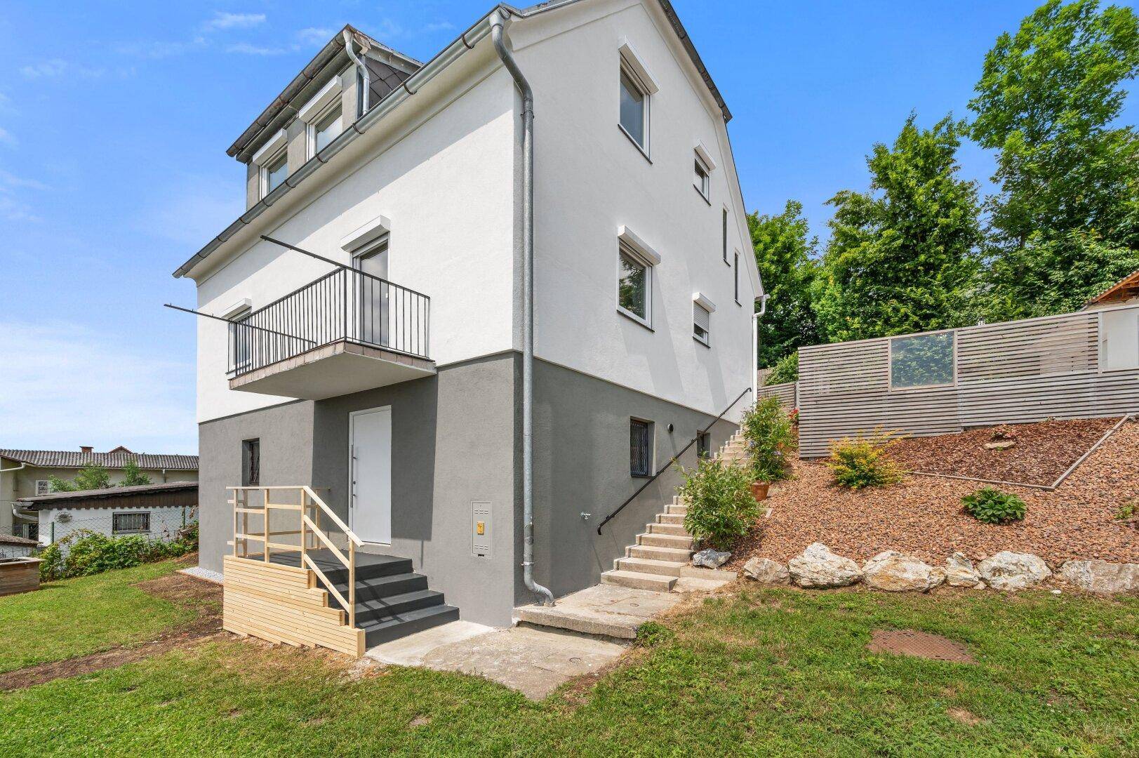 Seitenansicht des Hauses mit Außentreppe, Balkon und terrassiertem Gartenbereich.