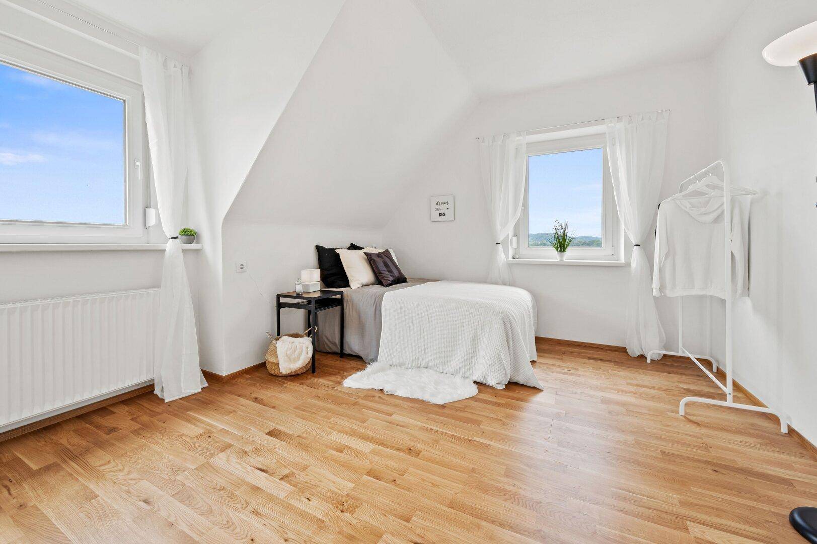 Helles Schlafzimmer im Dachgeschoss mit Holzboden, zwei Fenstern und Dachschrägen.