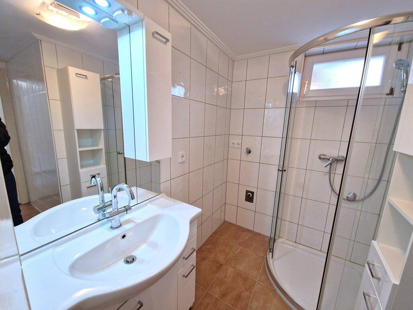 Badezimmer mit Waschtisch, Spiegelschrank und Eckduschkabine, hell gefliest.