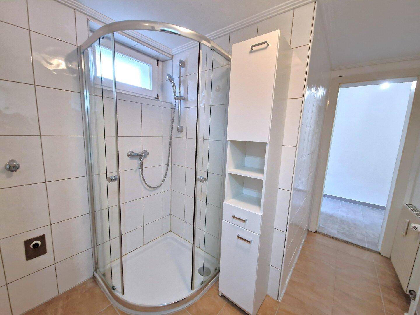 Badezimmer mit Eckduschkabine, weißen Wandfliesen und praktischem Hochschrank.