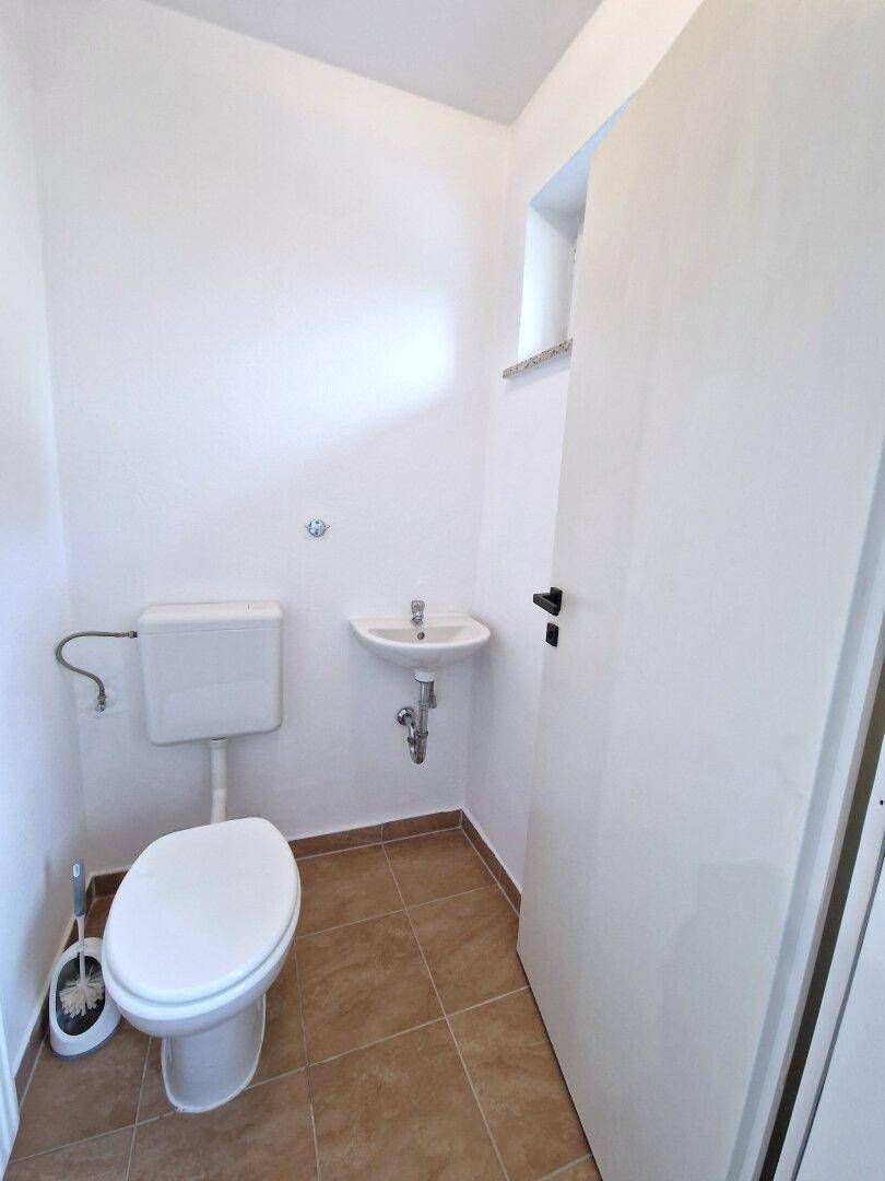 Gäste-WC mit Toilette, kleinem Waschbecken und Fenster, hell gefliest.