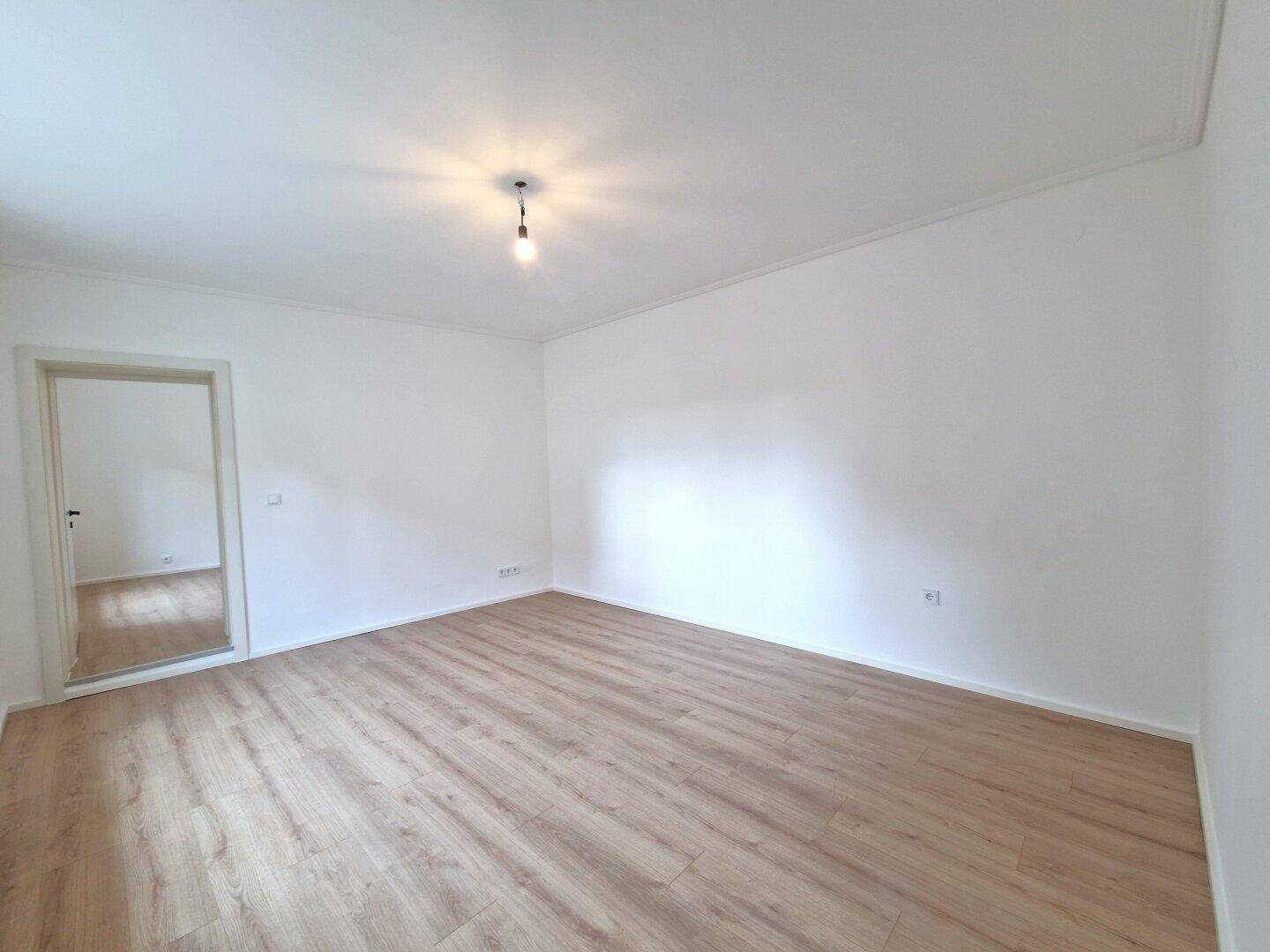 Leeres, helles Wohnzimmer mit Laminatboden und Zugang zu einem weiteren Raum.